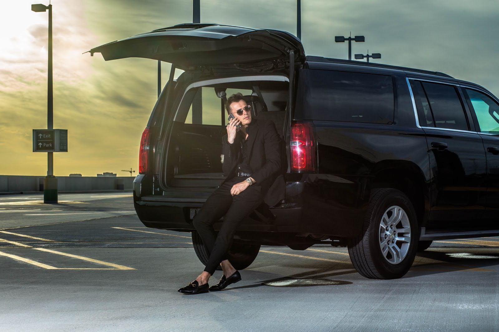 A professional man in a black suit talking on a phone while sitting in the open trunk of a luxury black SUV at an airport parking lot during sunset.