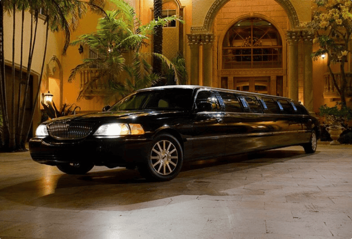 A long black luxury stretch limousine parked in front of a grand, lit-up mansion at night with palm trees in the background.